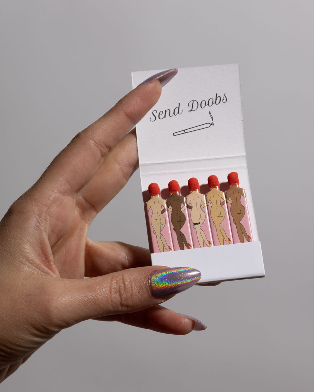 Woman with iridescent manicure holding a matchbook with 'send doobs' quote and matchsticks in the shape of women.